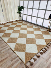 The RAMSHA HOME Beautiful Handmade Natural & Bleach Color Jute Braided Rug with Tassel (BR 031) features checkered tan and cream squares with tassels on two edges, displayed on a wooden floor near a window, plant, and cream curtains.