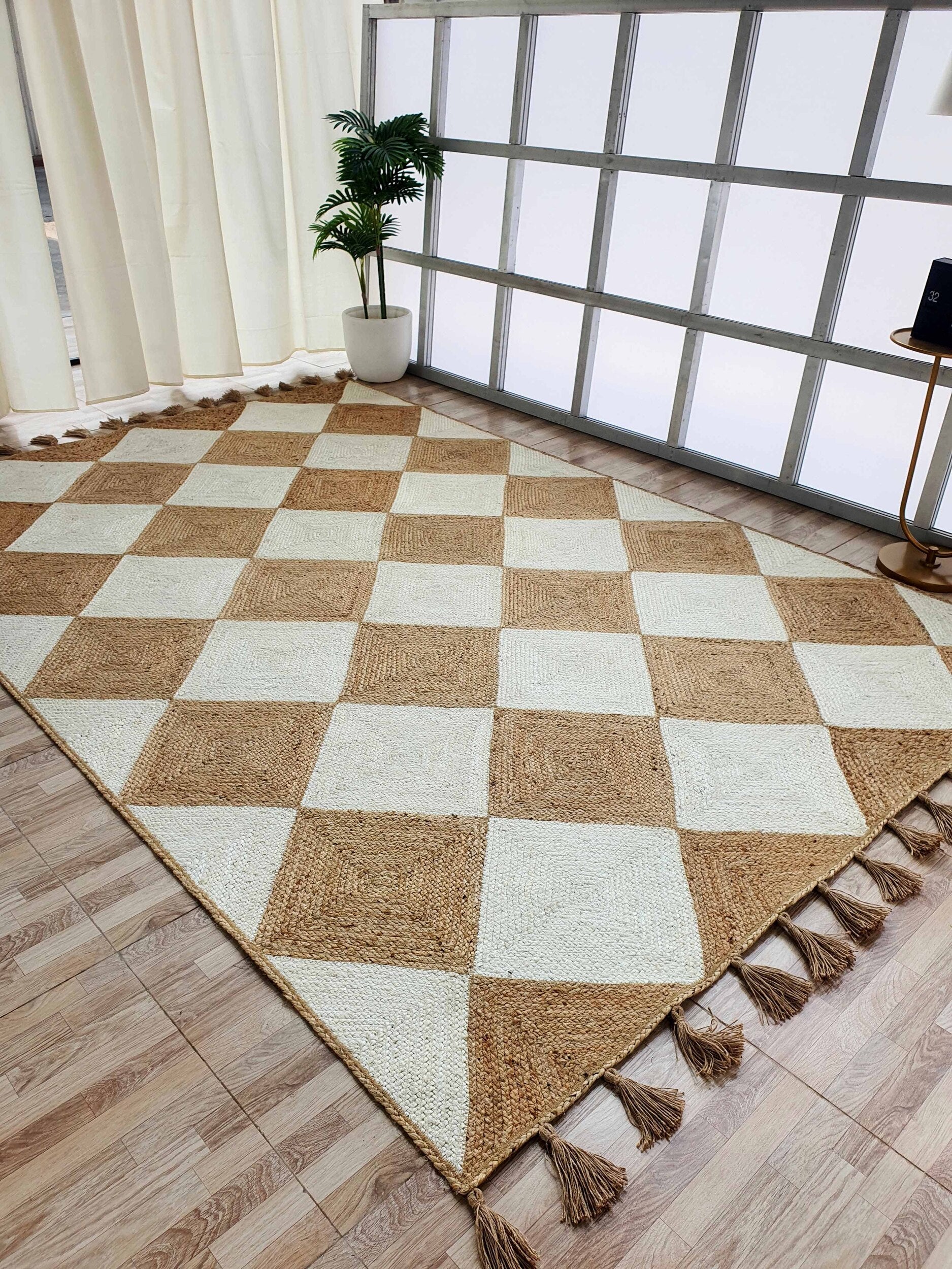 The RAMSHA HOME Beautiful Handmade Natural & Bleach Color Jute Braided Rug with Tassel (BR 031) features checkered tan and cream squares with tassels on two edges, displayed on a wooden floor near a window, plant, and cream curtains.