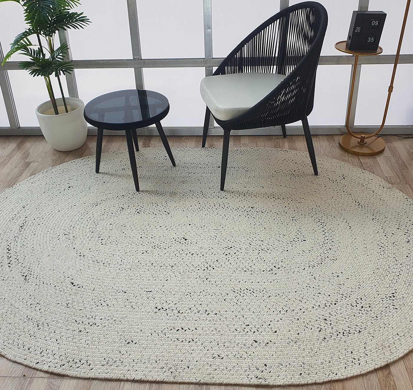 A modern sitting area with a black chair, small round black table, potted plant, and gold side table with a digital clock features the RAMSHA HOME Amazing Handmade White & Black Color Oval Wool Floor Braided Rug - BR 047 on a light wood floor by large windows.