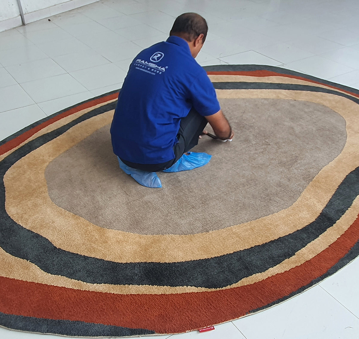 A man in a blue shirt and shoe covers kneels on the RAMSHA HOME Stunning Striking Multi Color Bedroom Decor Novara Rug - CMN 02 (200x300cm) in a tiled room, working on something atop its brown, orange, and black abstract rings.