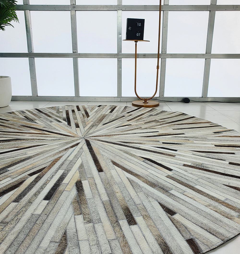 The RAMSHA HOME Home Decorative Leather Rug - LR 018, featuring a modern round design with a radiating geometric pattern in neutral tones, is placed on a white floor near a gold lamp and calendar by large frosted windows, with a white planter on the left.