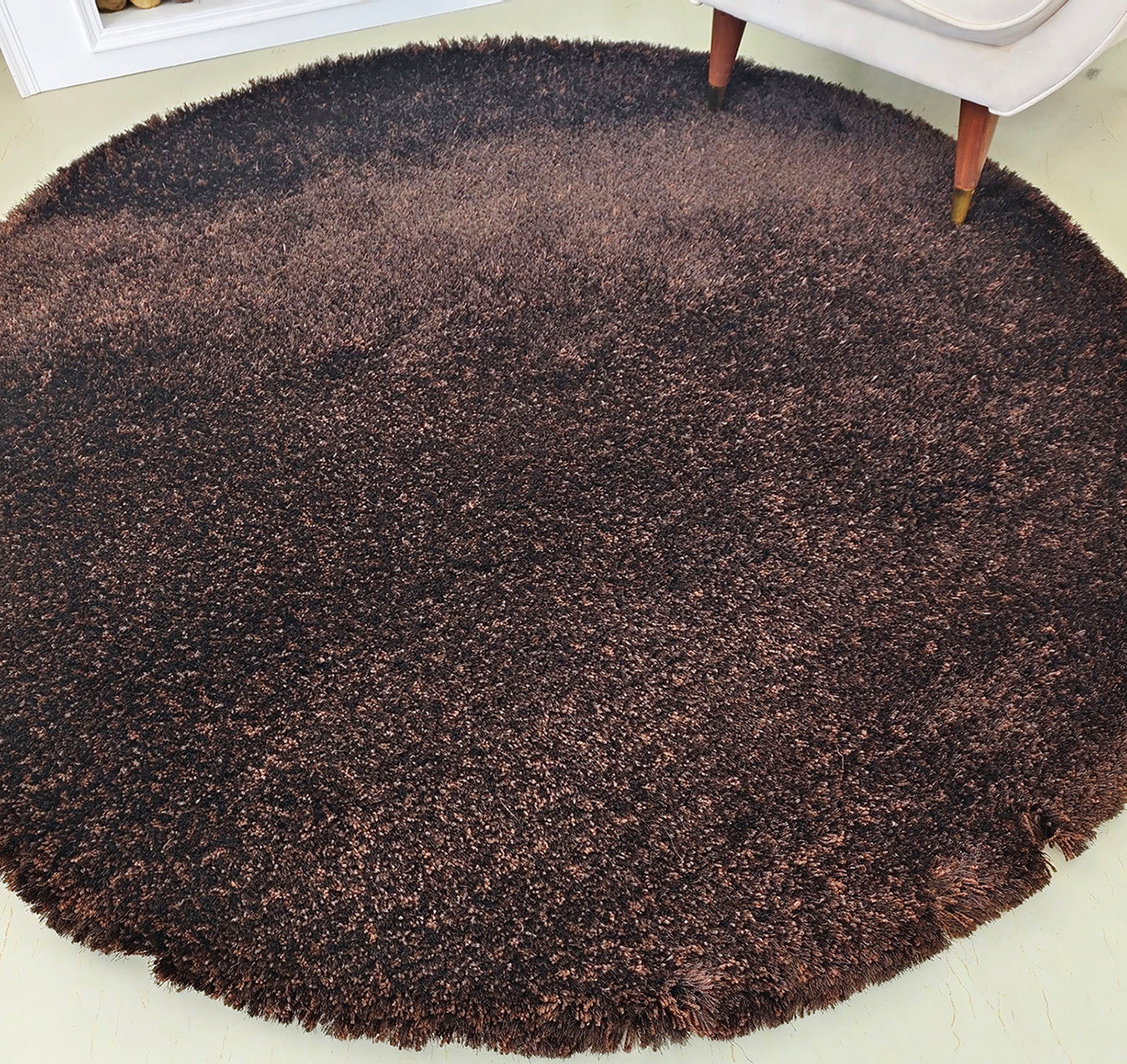 The RAMSHA HOME Fluffy Shaggy Round Shape Area Rug SG 04 in brown rests on a light floor. The bottom corner displays some fraying, and a light-colored chair leg is visible at the upper right edge.