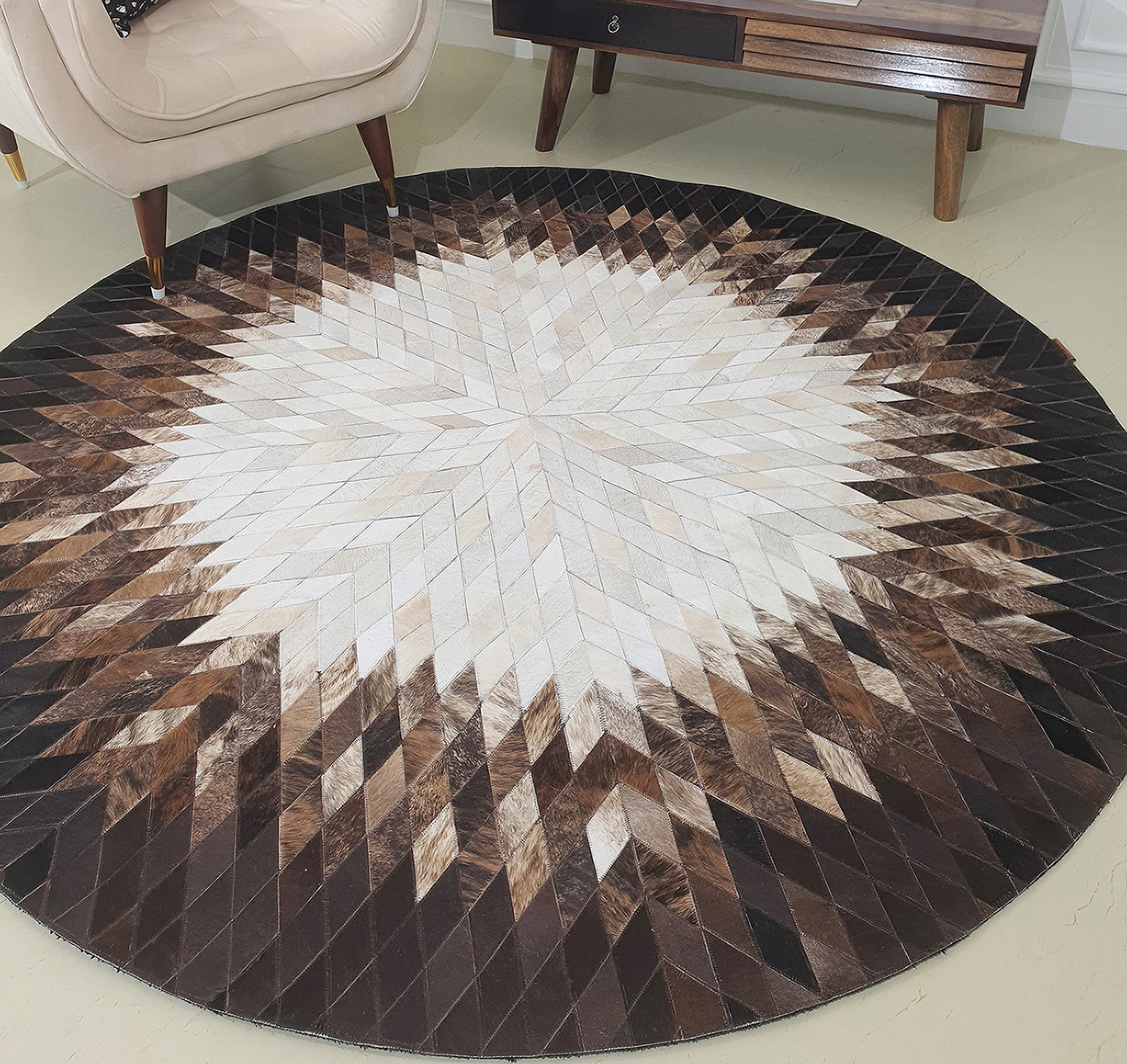 The RAMSHA HOME Multi Color Home Decor Round Leather Rug - LR 017 features a geometric starburst pattern with a light-to-dark gradient in white, tan, brown, and black. It’s styled near a beige chair and wooden console table.