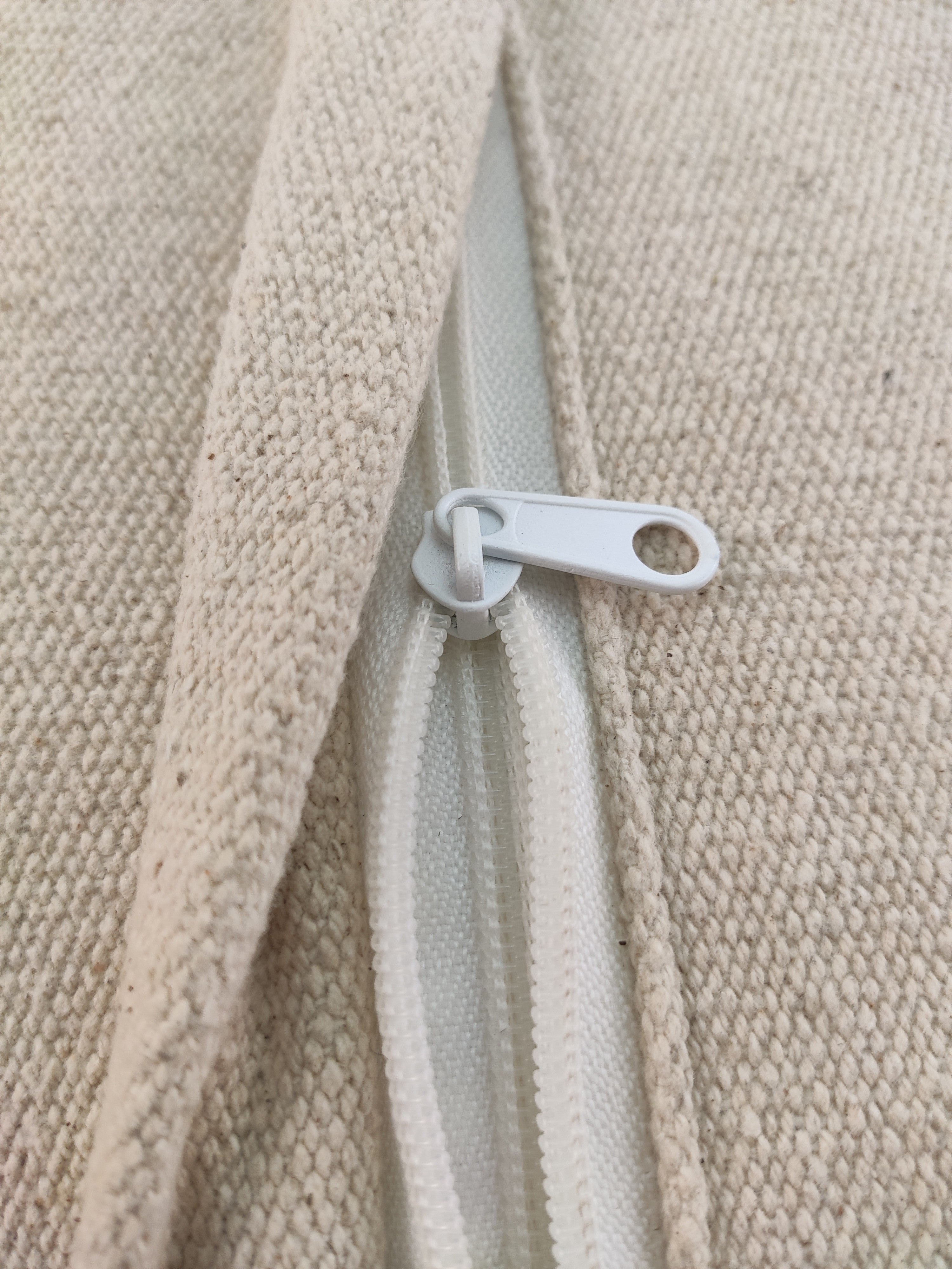 Close-up of a white zipper partially open on the beige textured fabric of the RAMSHA HOME Natural Wool Pouf With Leather Handle PF-045, showing the teeth and pull tab.