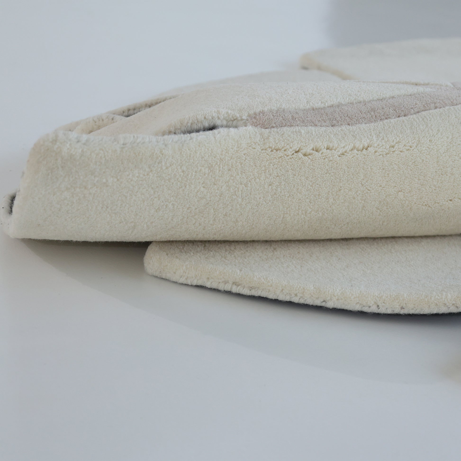 A close-up of the RAMSHA HOME Luxurious Brown & Beige Color Round Hand Tufted Rug (HTW 58, 200cm), folded to show its thick, textured, and plush fabric with subtle stitching details on a smooth white surface.