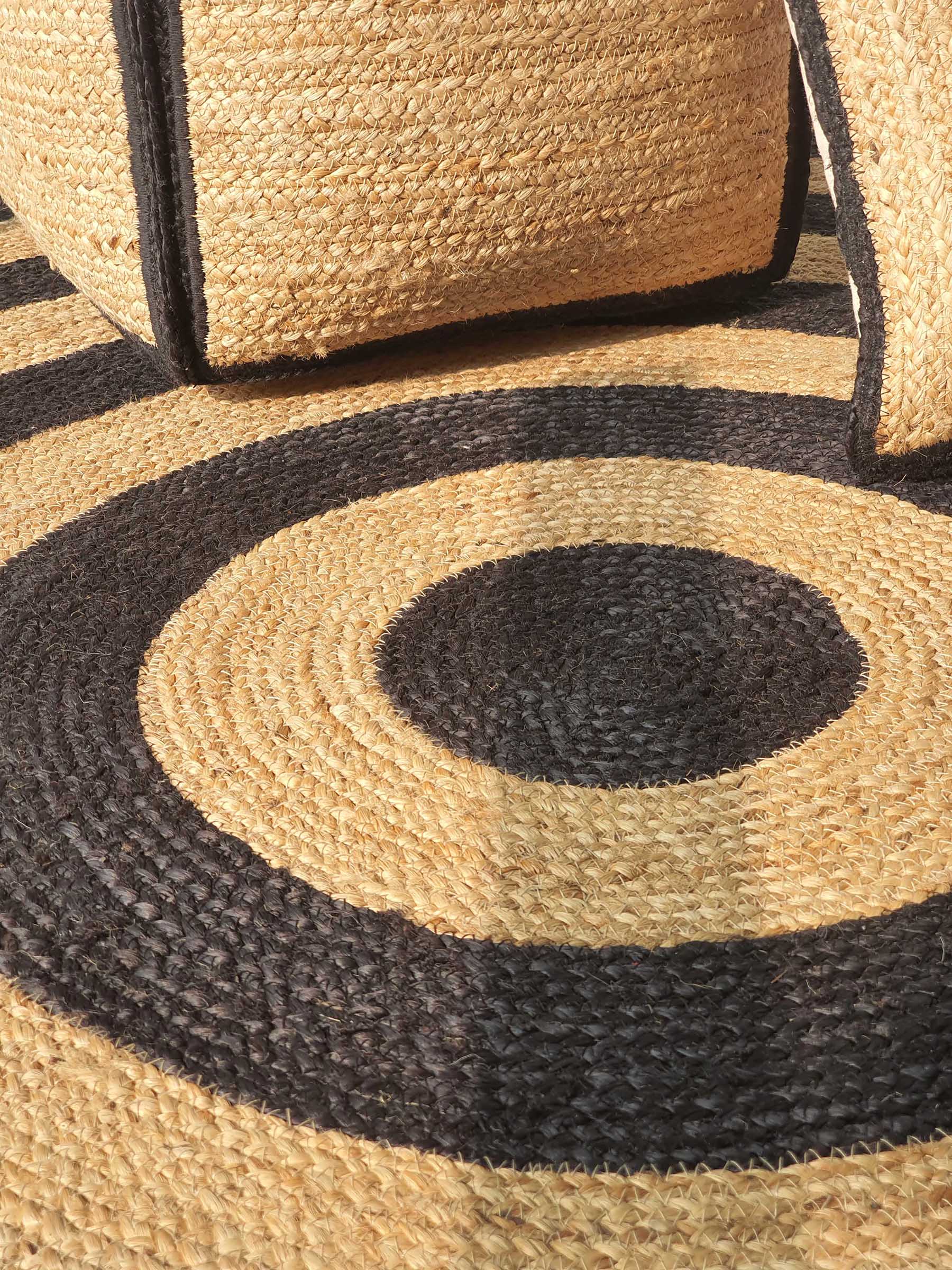 The RAMSHA HOME Handmade Braided Jute Floor Round Rug - BR 058 (200cm) features concentric black and natural circles, casting bold, graphic shadows in sunlight alongside a woven straw basket.