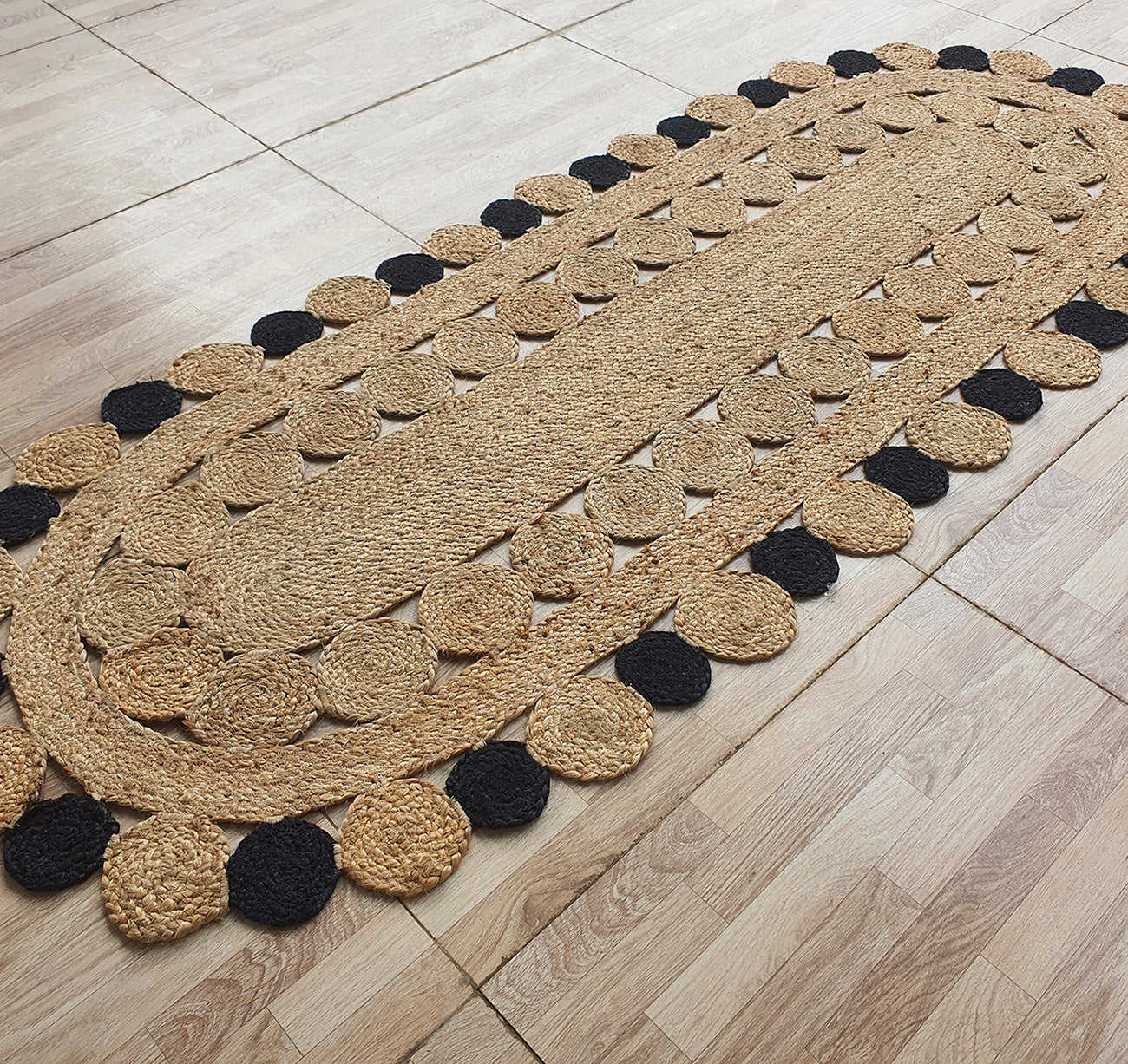 The RAMSHA HOME Oval Shaped Jute Braided Rug Black & Natural BR-033 features tan and black circular accents, adding a rustic and natural touch to a light wood floor with its braided jute texture.