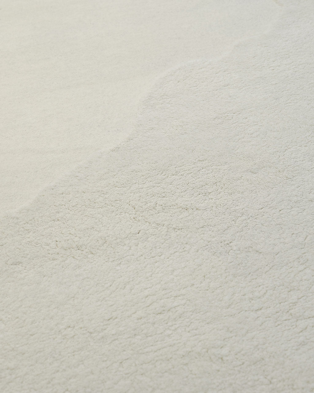 Close-up of the RAMSHA HOME Handmade Shape White Color Area Rug (HTW 101 B), showcasing its soft, off-white textured surface with subtle shading and an uneven edge that divides lighter and slightly darker areas.