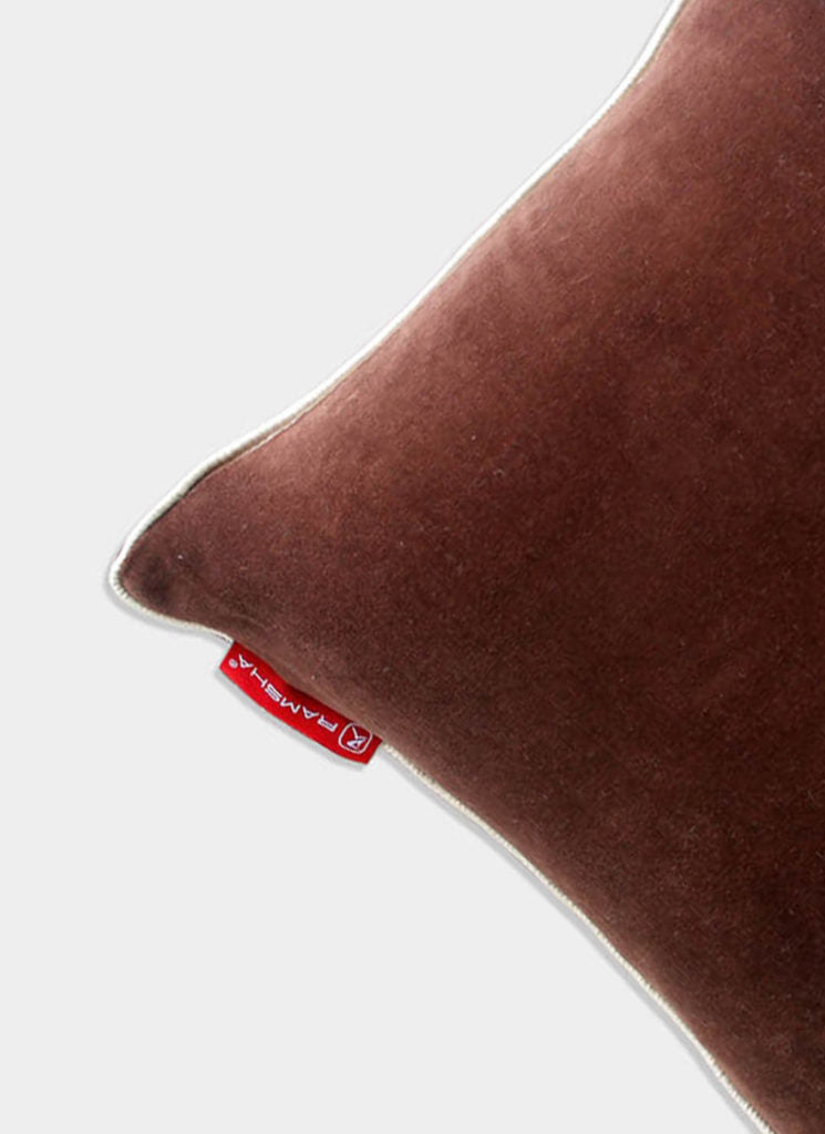 A close-up of the Polyester Cushion Plain With Filler cs-233 by RAMSHA HOME, a brown velvet plush cushion with white piping and a red fabric label, ideal for elegant home decor, set against a light gray background.