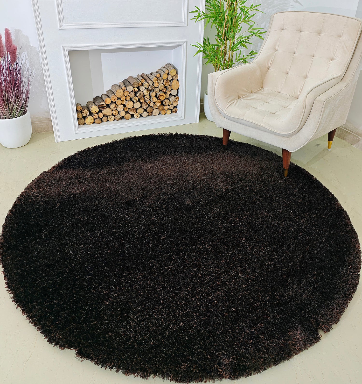 The RAMSHA HOME Fluffy Shaggy Round Shape Area Rug SG 04 in brown rests on a light floor next to a cream armchair, with a faux fireplace, stacked firewood, and a green potted plant behind.