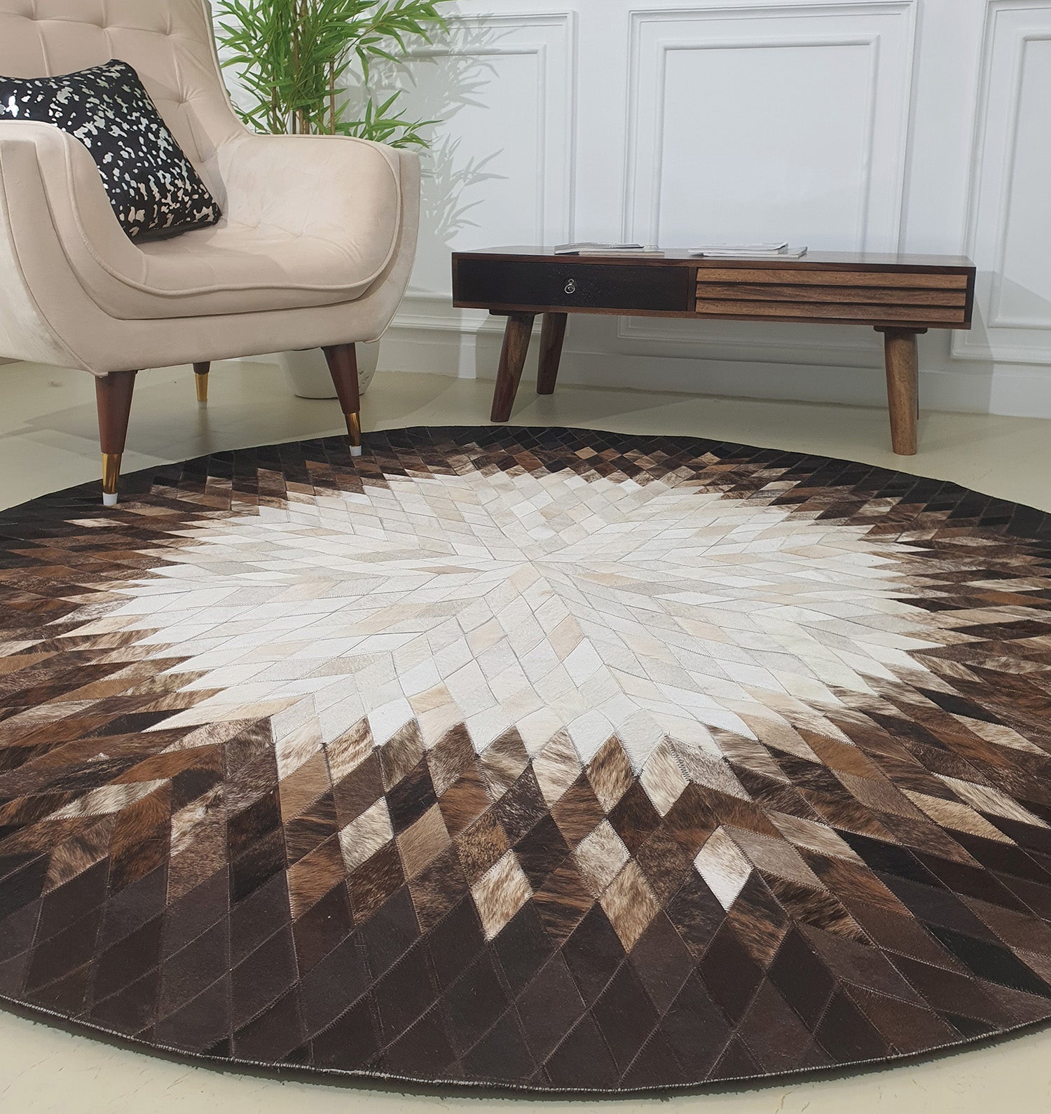 The RAMSHA HOME Multi Color Home Decor Round Leather Rug - LR 017, featuring a geometric pattern in brown, beige, and white, is placed near a modern beige armchair and a mid-century wooden table against a white paneled wall.