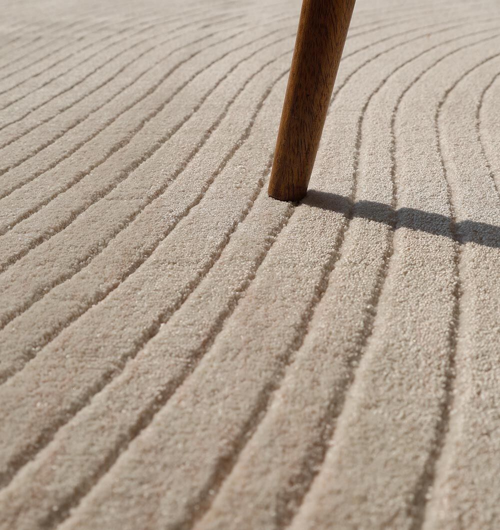 A close-up of the RAMSHA HOME Natural Handmade Area Rug - LKH-03 (300x400 cm) showcases its beige textured surface with curved, evenly spaced ridges, while a wooden furniture leg casts a shadow across this modern neutral rug.
