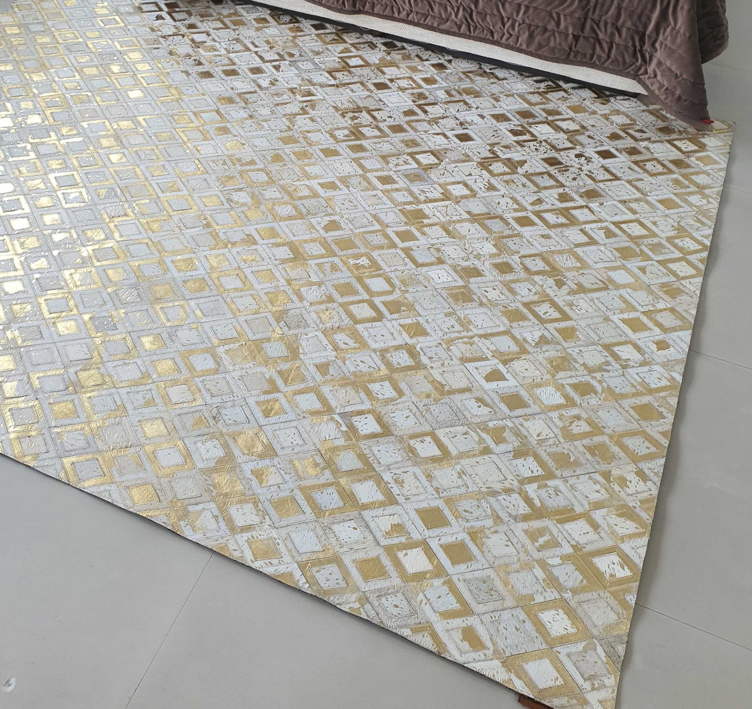 The RAMSHA HOME Handmade Diamond Patterned Leather Rug LR-010, featuring a gold and white geometric design, is displayed on a light floor beside a bed with a brown quilted cover.