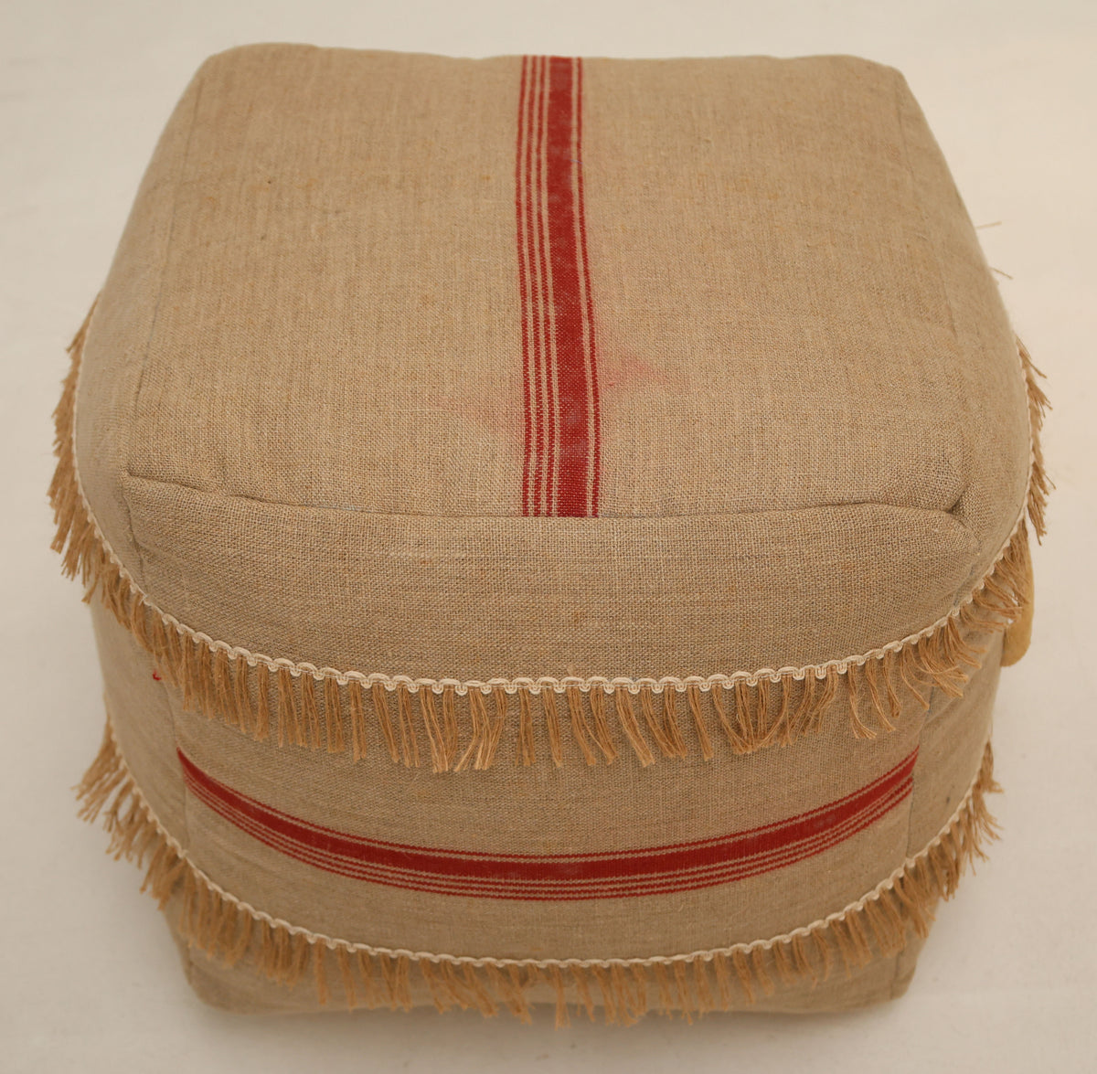 The Square Jute Floor Pouf with Fringes PF 192 by RAMSHA HOME, featuring a beige square design accented with red stripes and decorative fringe along its sides, sits on a light-colored floor and offers a stylish footrest or can be paired with a round pouf to enhance seating options and texture in your space.