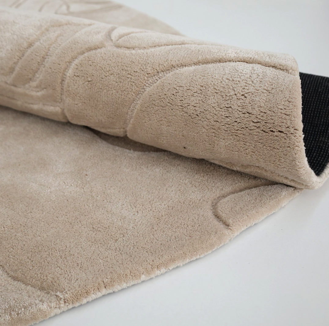 A close-up of the RAMSHA HOME Home Decor Handmade Area Rug - HTWV-156 shows its beige floral design and carved texture, partially rolled to display a black non-slip backing. The rug rests on a white surface, emphasizing its elegance.