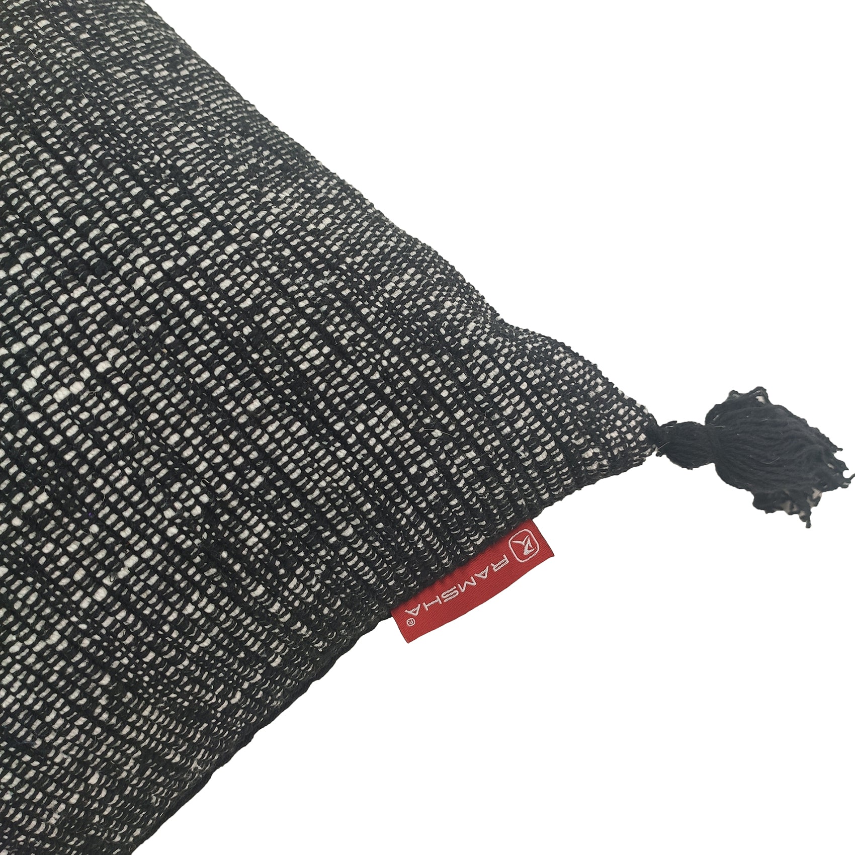 A close-up of a textured black and white Cotton Cushion With Fringes CS-133 by RAMSHA HOME, featuring a small black tassel at the corner, a red RAMSHA tag, and high-quality filler, displayed on a white background.