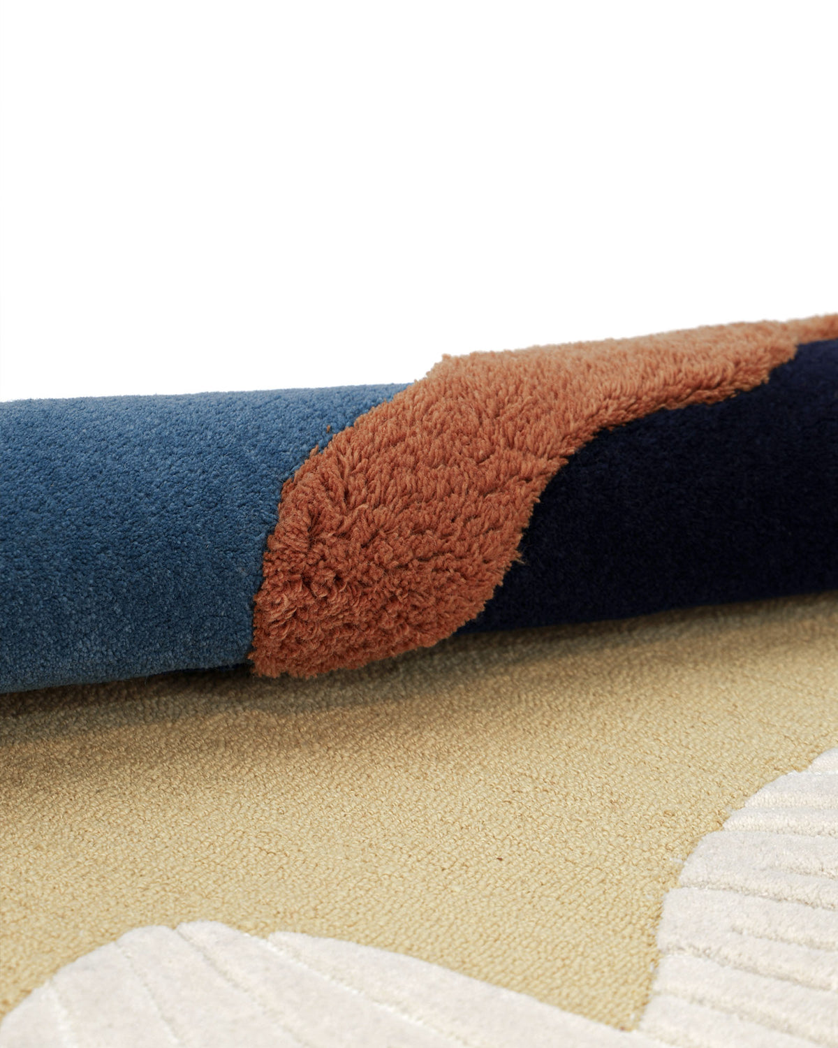 A close-up of the rolled-up RAMSHA HOME Handmade Wool & Cotton Abstract Round Area Rug HTW 125 (200cm) showcases hand-tufted, textured sections in blue, brown, and tan, with a distinctive white pattern on the flat part of this modern abstract design.