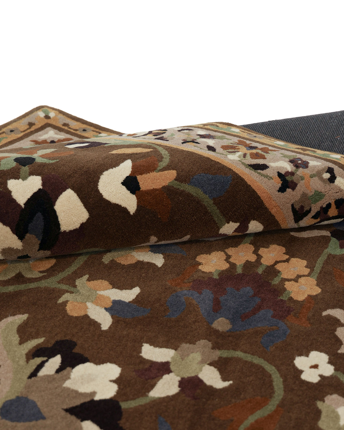 A close-up of the RAMSHA HOME Wool & Cotton Hand Tufted Rug HTW-07 reveals its traditional floral motifs in brown, cream, blue, and green hues. Partially rolled to display its thickness and texture, this hand-tufted rug highlights the intricate craftsmanship and superior quality that define the collection.
