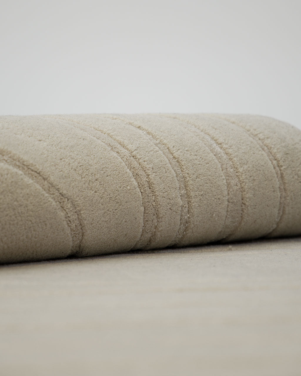 A close-up of the Hand Tufted Beige Color Decor Rug HTWV-108 by RAMSHA HOME, rolled up to showcase its textured surface with subtle, curved line patterns, placed on a matching beige floor. Crafted from hand tufted wool Tencel, this neutral area rug brings sophistication and comfort to contemporary spaces.