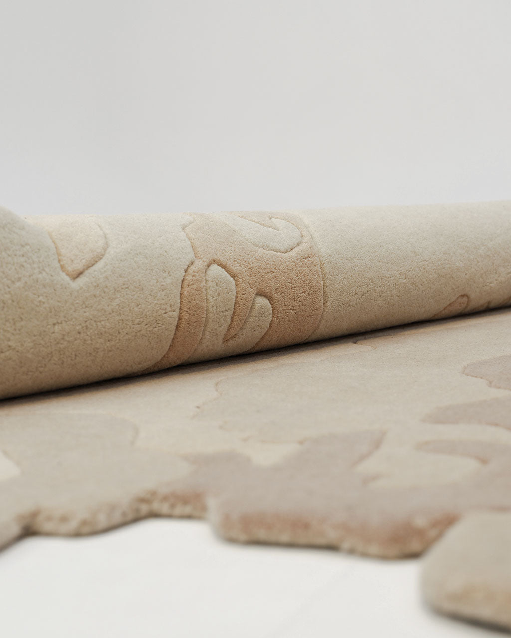 A close-up of the Handmade Shape Brown Color Area Rug - HTW 86 by RAMSHA HOME, showcasing its beige, textured abstract design with light brown patterns. Partially rolled on a white surface, this handtufted wool rug features an irregular shape that brings a modern touch to any space.