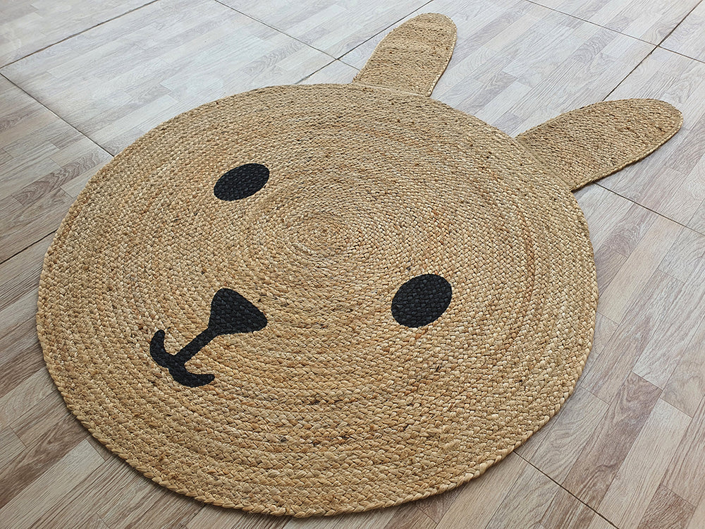The Amazing Handmade Jute Round Braided Rug - BR 078 by RAMSHA HOME, featuring a cute animal face with ear-shaped extensions, black eyes, nose, and mouth, is placed on a light wood floor.