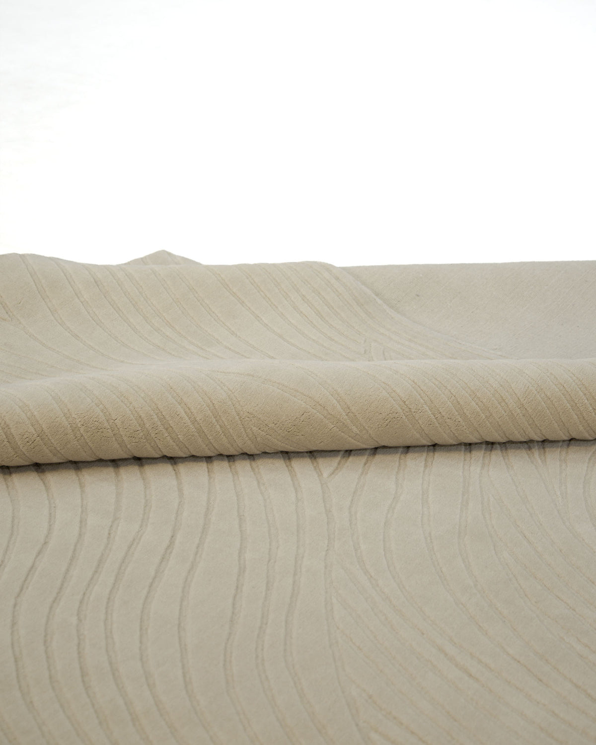 A close-up of the RAMSHA HOME Decorative Rectangle Handmade Beige Color 300x400 cm Area Rug (LKH-033) showcases its textured fabric with subtle, wavy lines. Part of the rug is rolled, highlighting its elegant pattern and plush, soft surface—evoking the luxurious feel of high-quality wool. The background remains plain and bright to emphasize the rug’s modern design.
