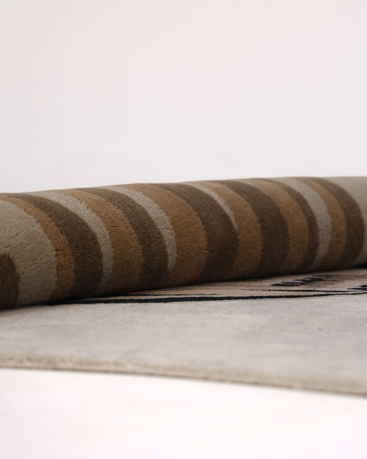A close-up view of the RAMSHA HOME Unshape Handmade Multi Color Rug HTW-82, featuring hand-tufted wool with brown and beige stripes, rolled up and resting on a flat, light-colored surface against a plain background.