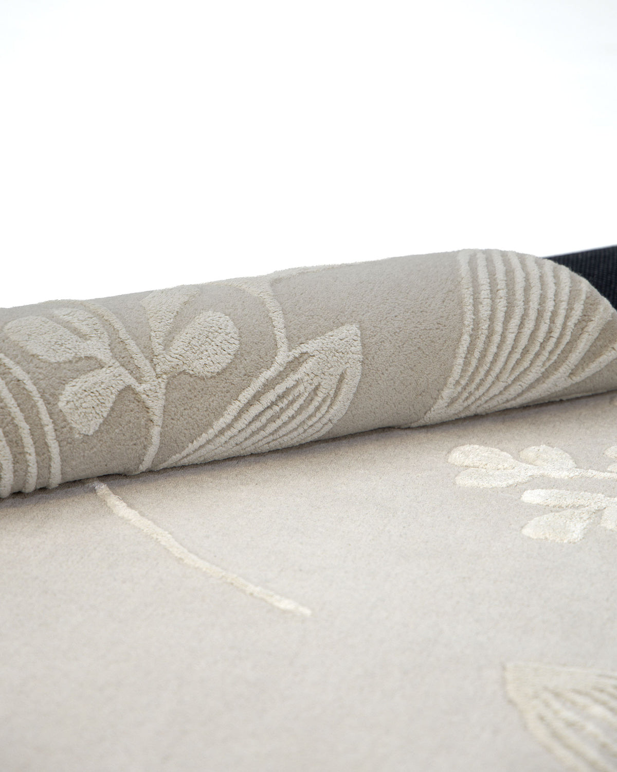 A close-up of the RAMSHA HOME Natural Beige & Ivory Handmade Area Rug (HTWV-146) shows its rolled edge featuring a subtle floral pattern in light cream and taupe tones, set against a plain background.