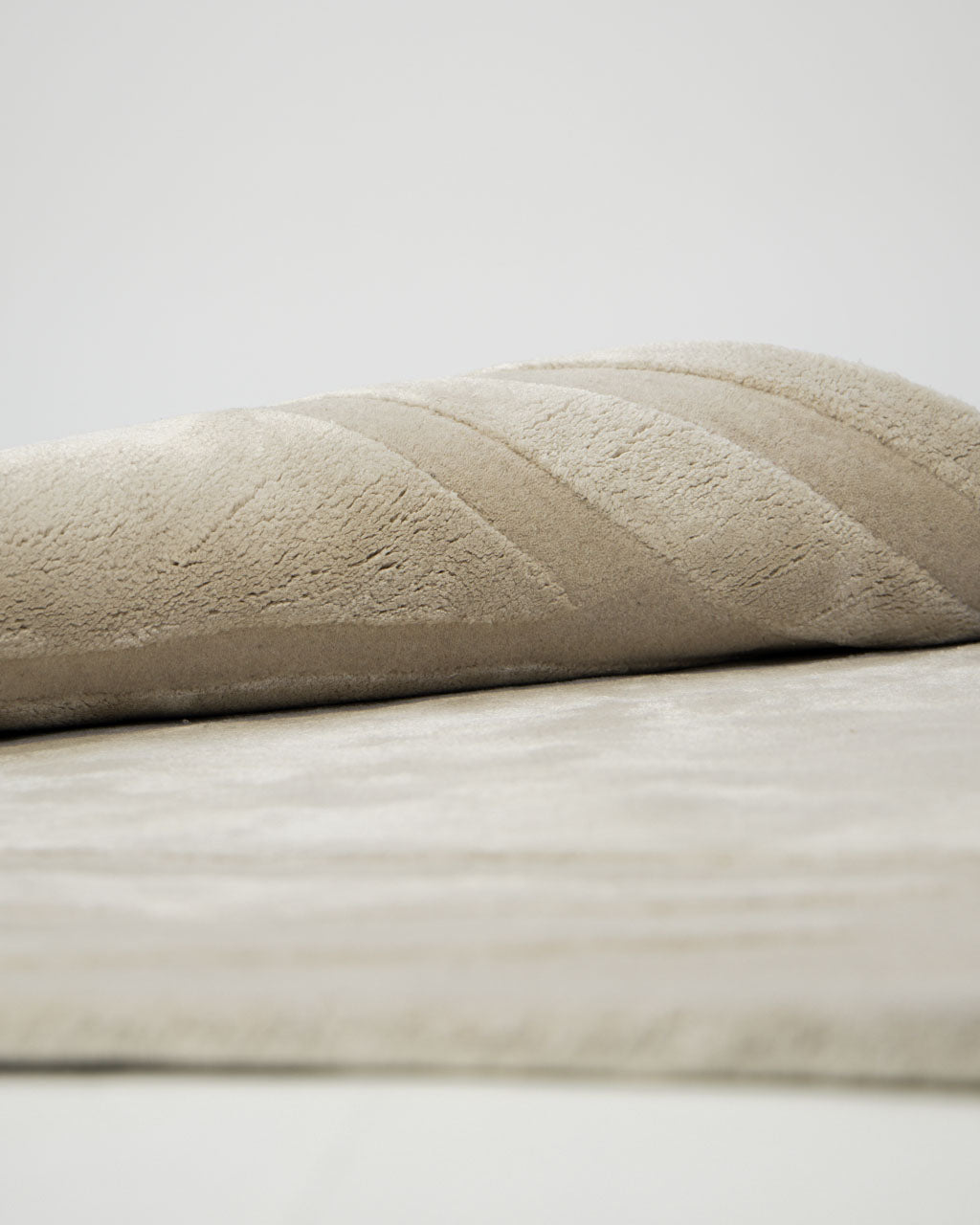 A close-up of the Handmade irregular 200x300 cm Rug HTWV-198 by RAMSHA HOME reveals its soft cream and beige hues, with subtle diagonal lines, displayed folded on a light surface against a plain background—evoking a gentle, inviting texture.