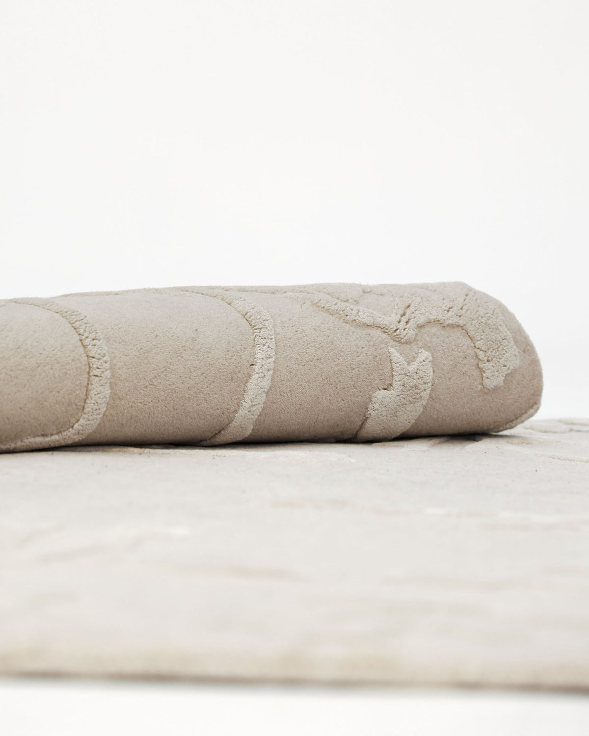 A close-up side view of the RAMSHA HOME Decorative Handmade Area Rug HTWV-118, featuring a light beige or cream-colored hand-tufted wool blend with subtle textured patterns, partially rolled against a plain white background.