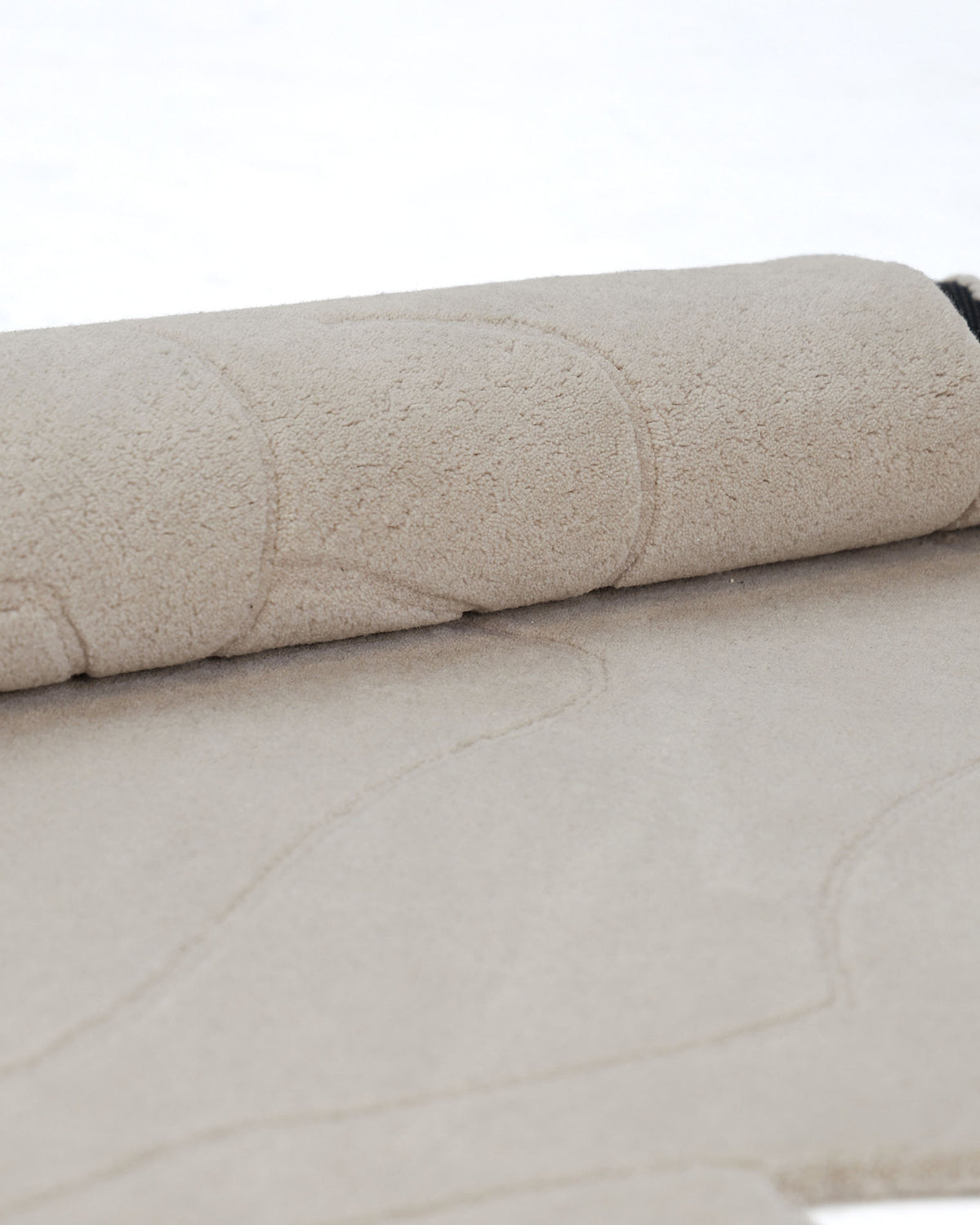 A close-up of the RAMSHA HOME Handmade Rose Shape Rug HTW 106 in light beige, partially rolled up to reveal its delicate, wavy line patterns. The rug is set against a clean, white background.