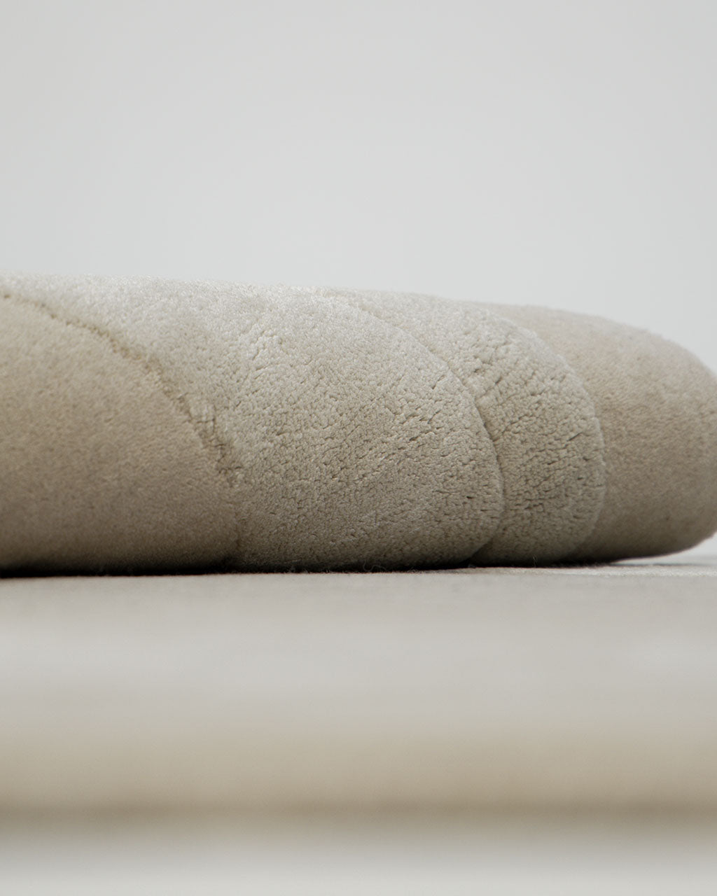 A close-up of a soft, light beige rolled towel or blanket rests on a smooth surface, accentuating its plush, textured fabric and gentle curves, set atop the RAMSHA HOME Handmade irregular 200x300 cm Rug HTWV-191 in subtle cream and beige tones.