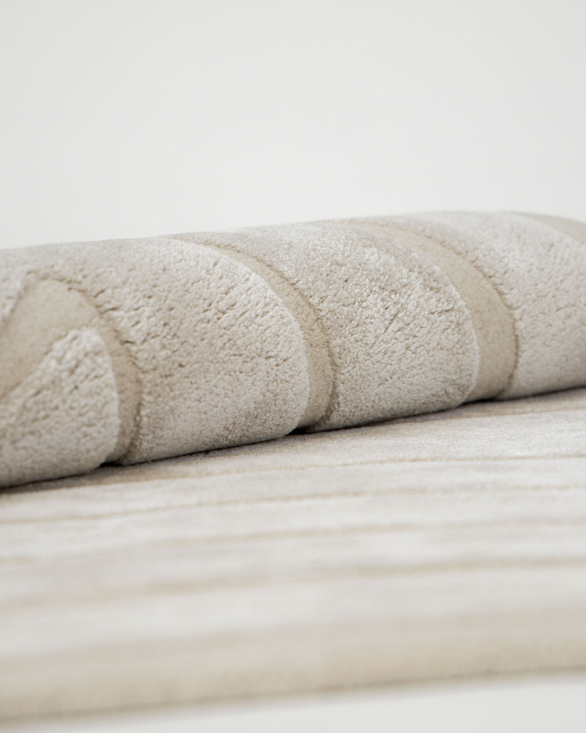 A close-up of the RAMSHA HOME Luxurious Off-White Hand-Tufted Rug - HTWV 111 (200x300cm) shows its plush, soft texture and subtle curved patterns, rolled up and resting on a matching flat rug surface against a plain background.