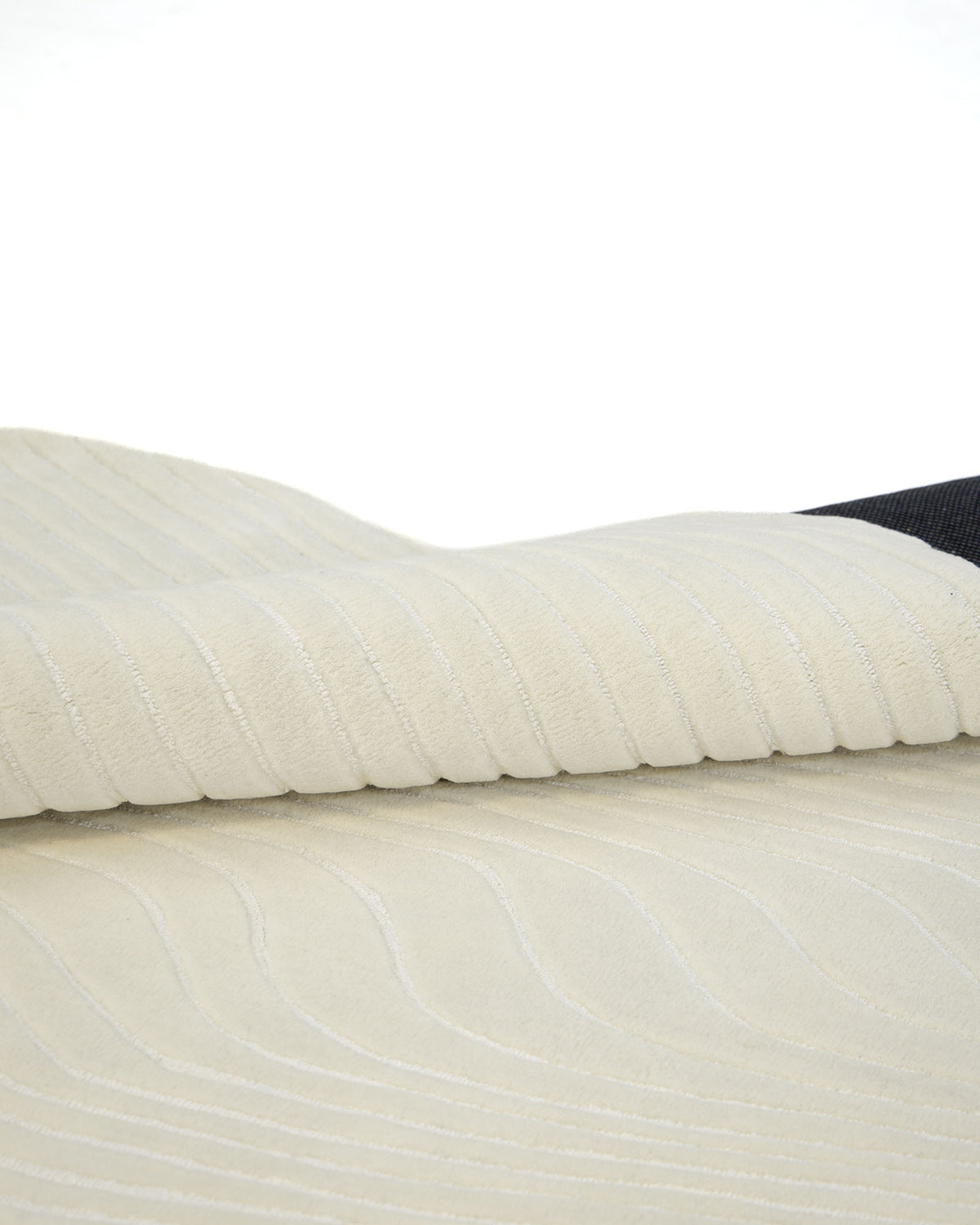 A close-up of the RAMSHA HOME Hand Tufted Multi Color Decor Rug HTWV-101, showcasing its folded cream-toned surface with a textured, wavy, and ribbed pattern—evoking the look of an irregular wool rug—as it partially drapes over a dark background.