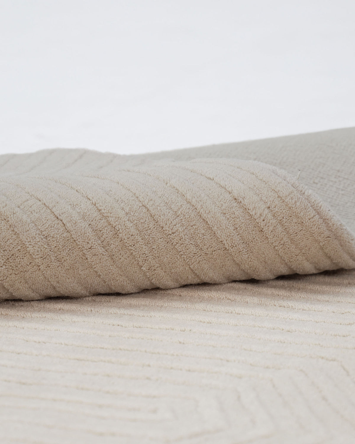A close-up of the RAMSHA HOME Home Decor Handmade Area Rug - LKH-014 (200x300 cm) in beige, partially rolled on a matching surface, highlights its soft, ribbed texture reminiscent of ivory wool.