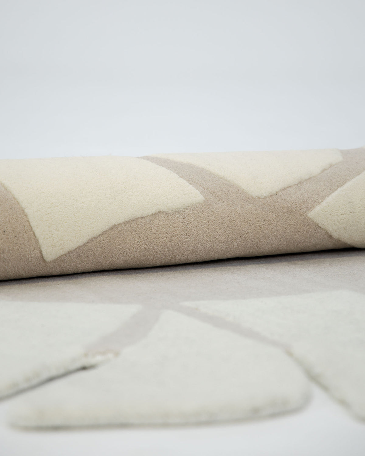 A close-up of the RAMSHA HOME Luxurious Brown & Beige Color Round Hand Tufted Rug - HTW 58 (200cm) showcases its abstract shapes in rolled form, resting on a flat surface with a matching pattern in the foreground. The plain white background accentuates the rug’s intricate design.