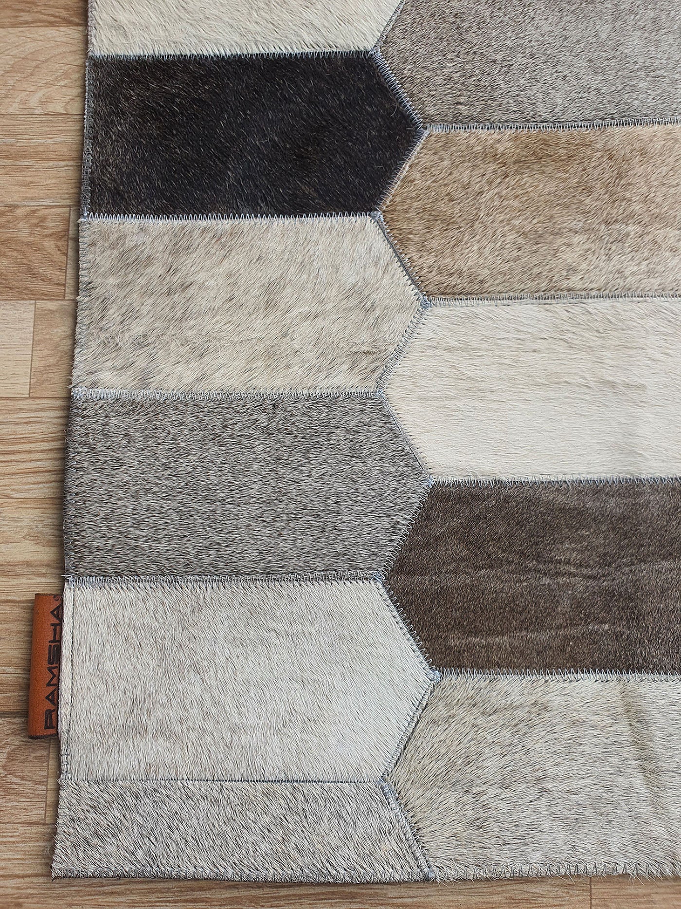 A close-up of the Multi Color Home Decor Leather Rug - LR 011 by RAMSHA HOME, featuring a hexagon geometric pattern in brown, beige, gray, and cream tones on a wooden floor. A branded tag is visible on the edge of this stylish leather patchwork rug.