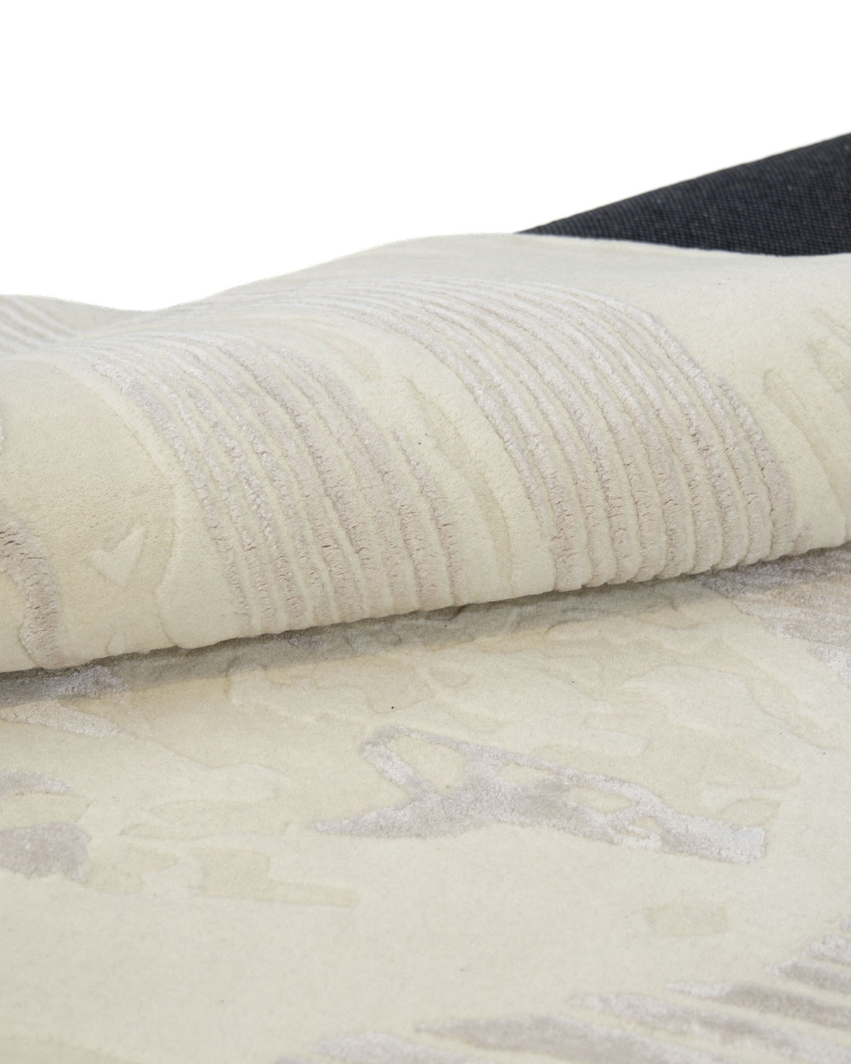 A close-up of the RAMSHA HOME Abstract Beige Handcrafted Rug – Neutral Textured Modern Area Rug (HTWV 205) shows its light-colored, textured surface with subtle raised patterns, while a folded corner reveals the dark backing underneath.