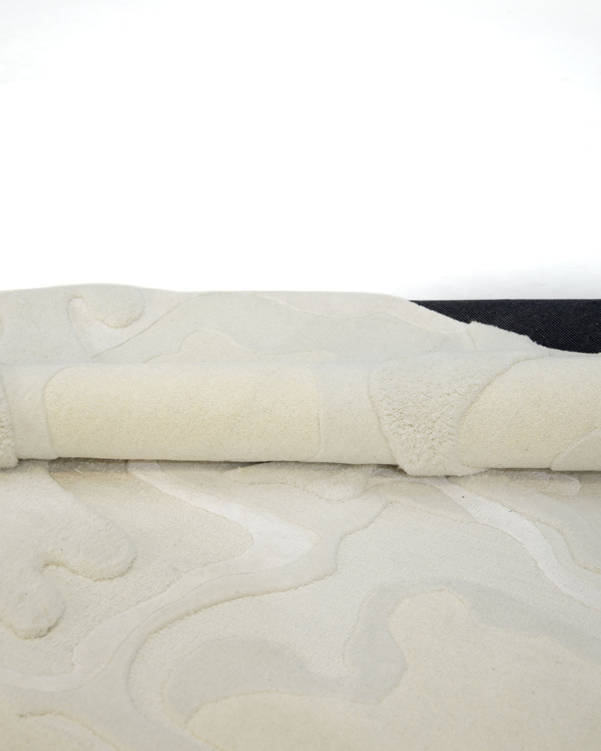 A close-up of the Handcrafted Natural White Area Rug – HTWV-129 by RAMSHA HOME reveals its modern abstract design in cream tones with beige patterns, partially rolled up on a white surface. This handtufted rug elegantly highlights its unique artistry and soft texture.