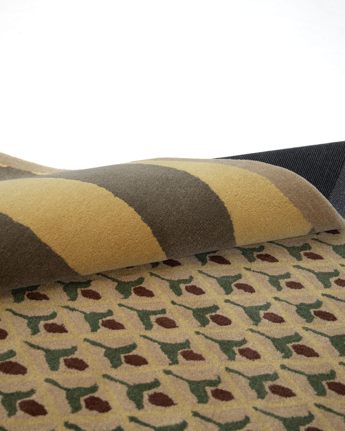 A close-up of the RAMSHA HOME Modern Mustard & Grey Wool Carpet HTW-30, hand-tufted and partially folded to reveal a striped beige and olive backing, while the front displays a floral pattern with brown, yellow, and green accents.
