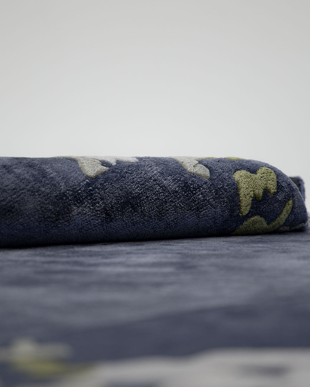 A close-up view of a rolled, dark blue towel featuring green and white patterns, placed on the RAMSHA HOME Navy Blue Viscose Shaped Handmade Carpet 200x300 - HTV-23, set against a plain light background.