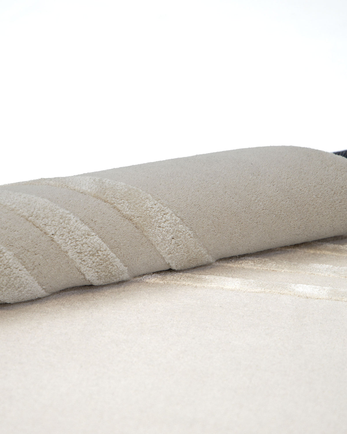 A close-up of the Hand-Tufted Carpet For Home And Office HTWV-70 by RAMSHA HOME showcases its rolled-up form, highlighting the beige hue, visible texture, and subtle striped patterns as it rests on a flat matching surface—perfectly capturing the understated elegance of a minimalist wool rug.