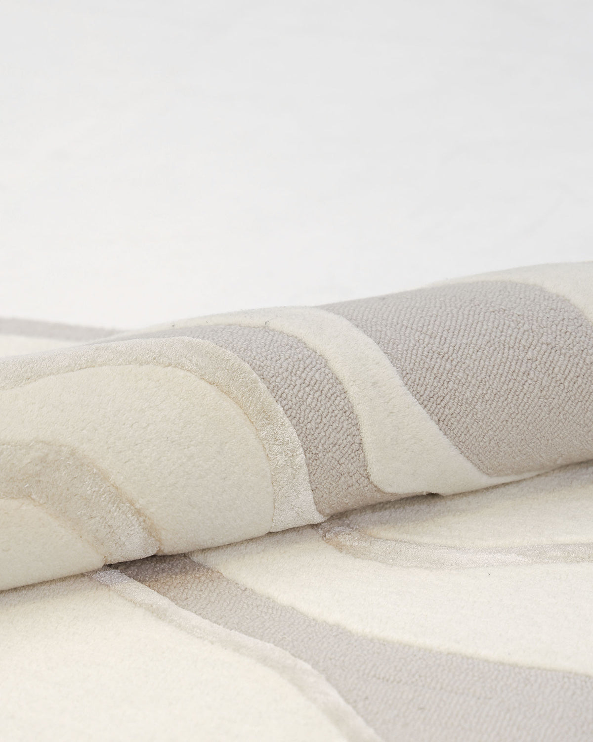 A close-up of the RAMSHA HOME Handcrafted Living Room Area Rug – HTWV-127 shows its beige and cream tones with elegant undulating wave patterns, partially rolled over on a light backdrop. The handtufted texture and raised design details are clearly visible, making this neutral area rug an inviting addition to any space.