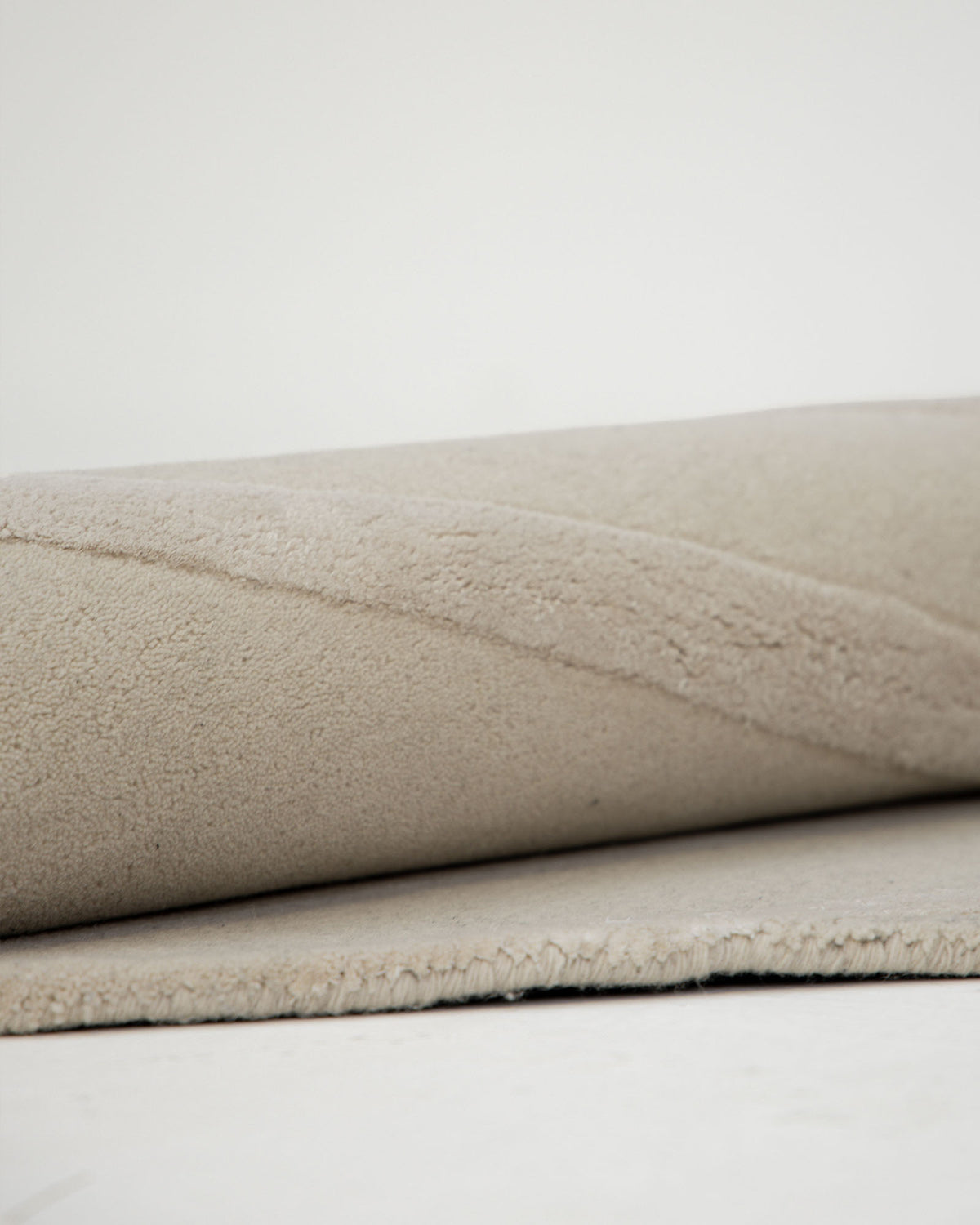 A close-up of the RAMSHA HOME Multi Color Handmade Area Rug - HTWV-136 (200x300 cm) rolled up on a white surface, highlighting its soft, hand-tufted wool texture and subtle wavy pattern.