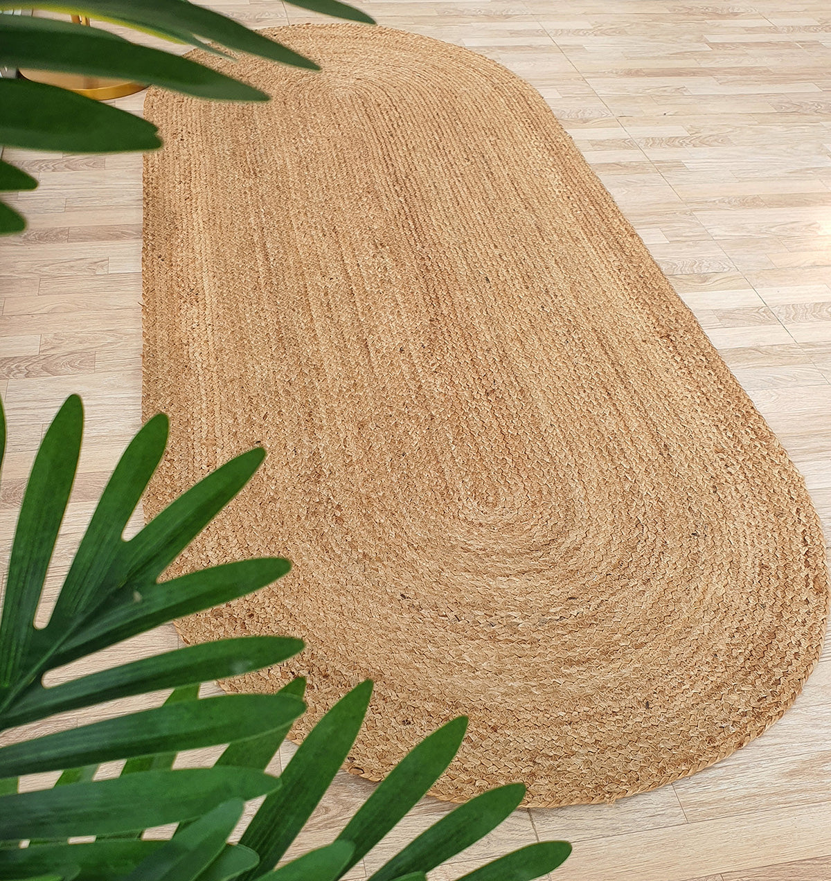 The Handmade Natural Color Oval Shape Braided Rug - BR 036 by RAMSHA HOME rests on a light wood floor, edged by green indoor plants. This jute rug infuses the space with organic texture and warmth.
