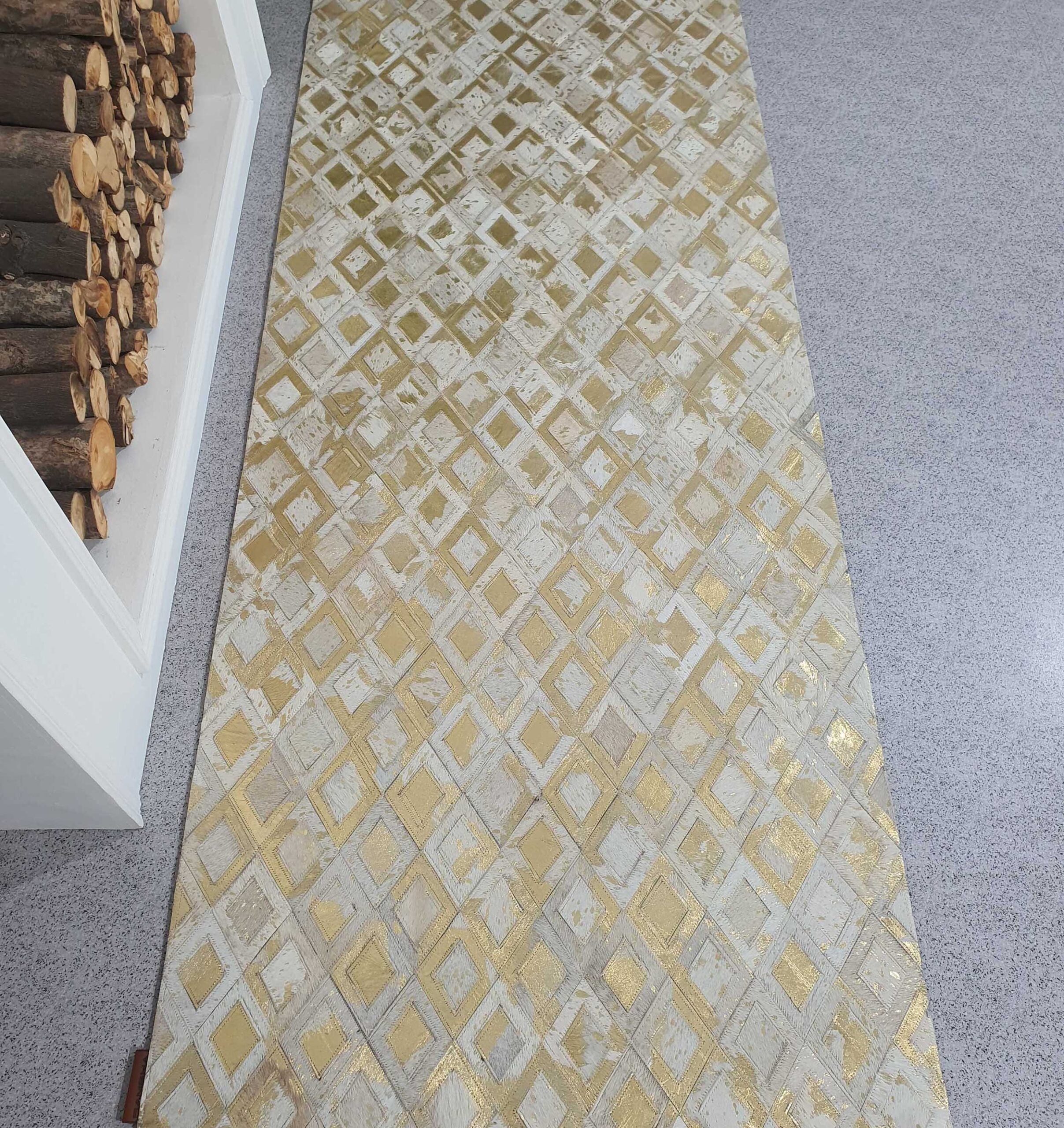 The RAMSHA HOME Handmade Diamond Patterned Leather Rug LR-010, featuring a gold and white geometric design, sits on a gray speckled floor beside a fireplace stacked with chopped firewood.