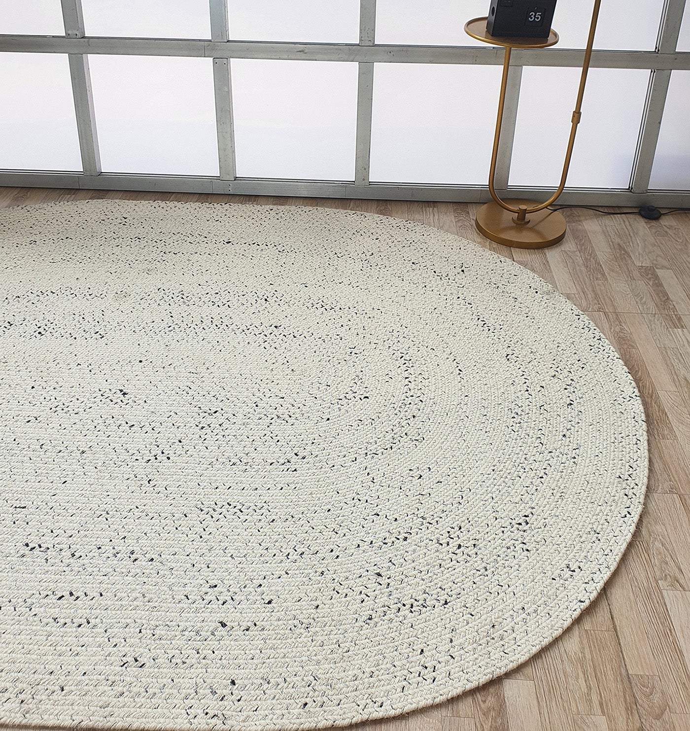 The Amazing Handmade White & Black Color Oval Wool Floor Braided Rug by RAMSHA HOME rests on a light wood floor near a modern gold side table with a small black object, set before tall frosted metal-framed windows.