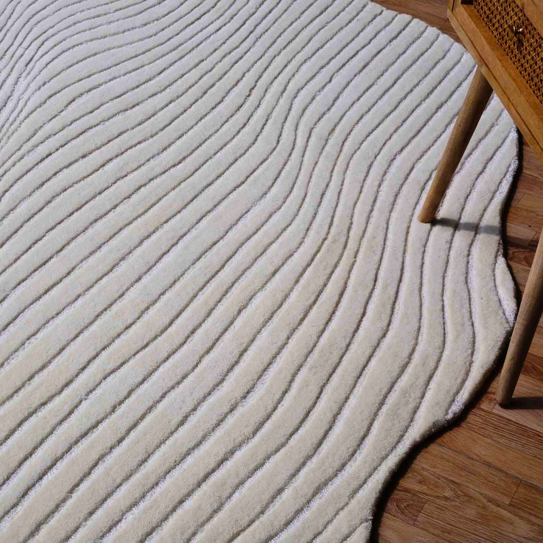 The RAMSHA HOME Hand Tufted Multi Color Decor Rug HTWV-101, featuring a cream hue and textured wavy raised lines, is placed on a wooden floor beside the leg of a wooden chair with a woven seat.