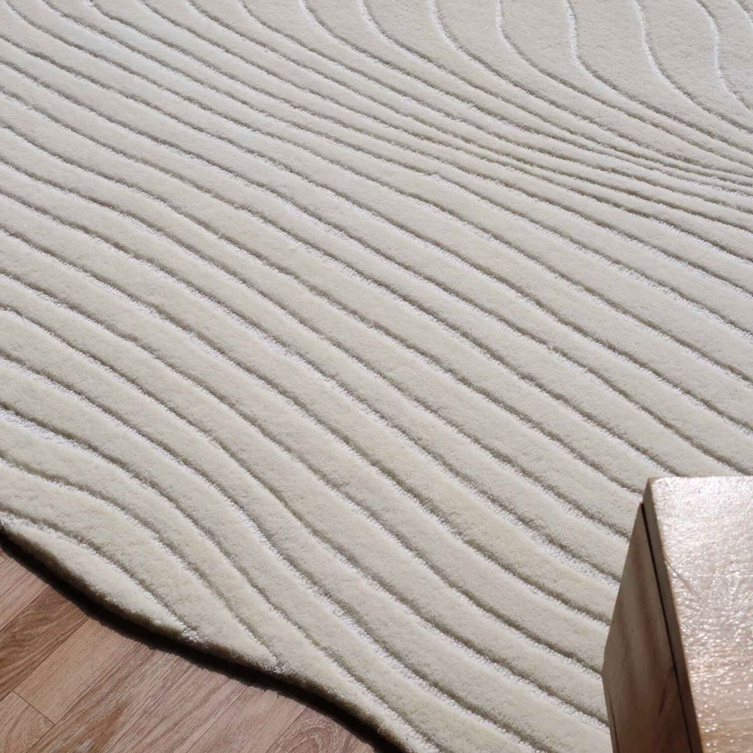 The Hand Tufted Multi Color Decor Rug HTWV-101 by RAMSHA HOME, an irregular cream wool rug with wavy raised lines, rests on a wooden floor. A light wood table’s corner appears in the lower right of the image.