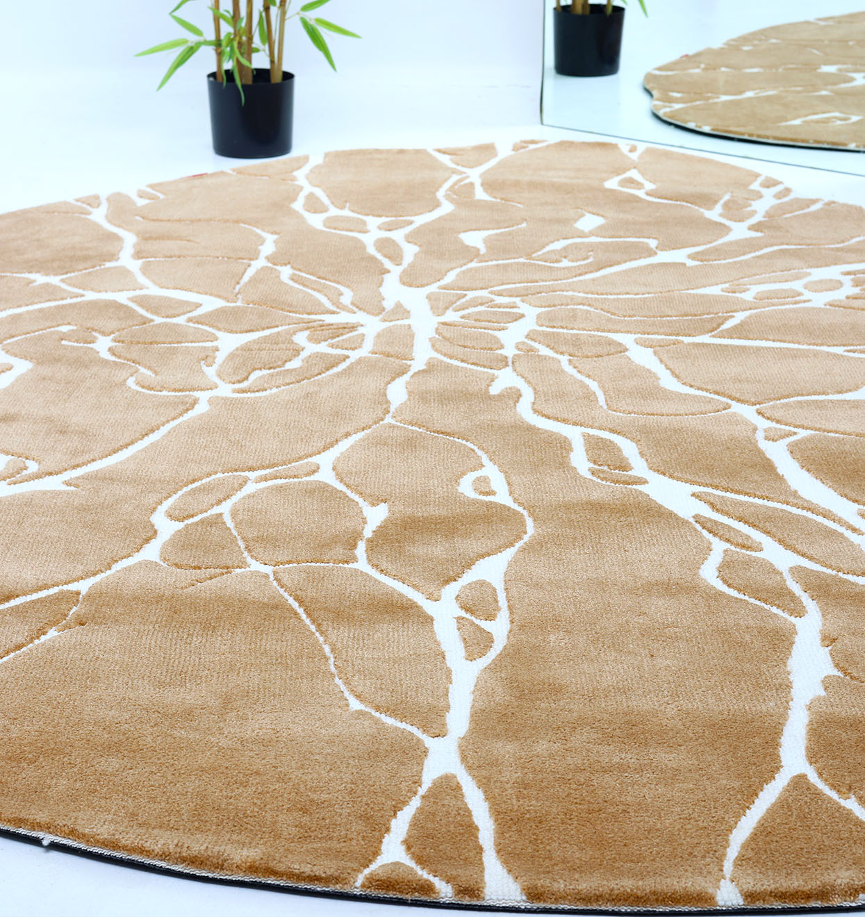 The Novara Shape Rug Multi Color CMN-13 by RAMSHA HOME, with beige and white abstract patterns, lies on the floor. In the background, two black plant pots hold green bamboo beside a circular mirror reflecting the plants and rug.